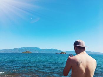 Rear view of shirtless man photographing sea against clear sky