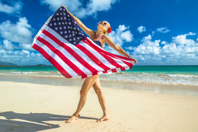 Happy woman holding american flag while standing at beach against blue sky