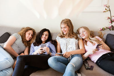 Happy teenage girls using smart phone while reclining on sofa in living room