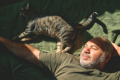 Top view of bearded middle-aged man lying on a bed and playing with his gray tabby cat.