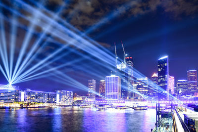 Illuminated bridge over river by buildings against sky at night