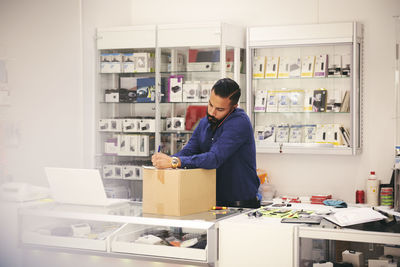 Salesman talking on mobile phone while writing on box at counter in electronics store