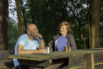Tired man and woman sitting o camping bench and table driking and having a talk sorrounded by nature