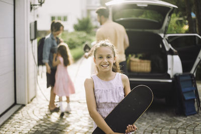 Smiling girl holding skateboard with family in background