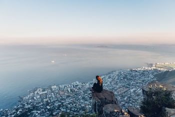 Side view of young woman looking at view while sitting on mountain against sky during sunset