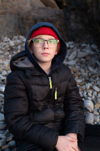 Tween boy sitting on beach of stones wearing glasses watching sunset