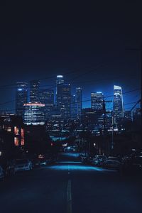 Illuminated city street by buildings against sky at night