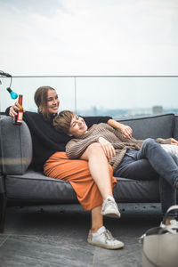 Smiling woman leaning on female friend with beer over sofa at building terrace