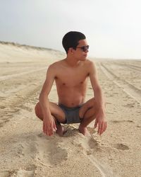 Shirtless young man crouching on sand at beach against sky