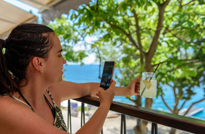 Young woman using mobile phone, taking photos of jar of fresh lemonade in beach bar
