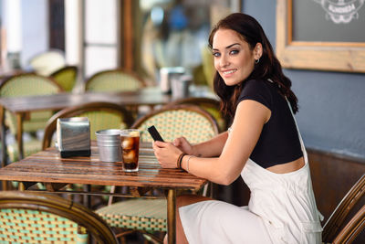 Portrait of smiling young woman using phone while sitting at sidewalk cafe