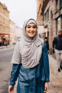 Portrait of smiling young muslim woman wearing hijab walking on sidewalk in city