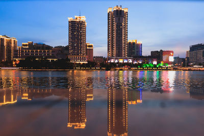 Illuminated buildings by river against sky in city