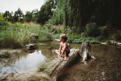 Young boy sitting on log in water in summer looking thoughtful