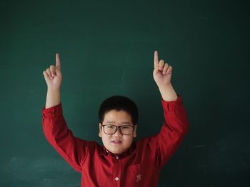 Portrait of boy with arms raised standing against blackboard in classroom