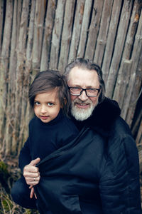 Grandson in a blue hat and boots with a grandfather in a sheepskin coat stand at a wooden house shed