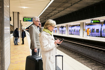 Senior couple using smart phones while standing with luggage at illuminated subway station