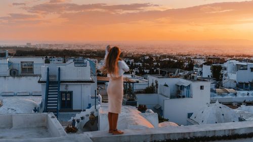 Rear view of woman standing by railing against sky during sunset