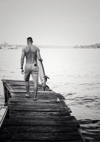 Full length of shirtless man walking with surfboard on jetty over sea against clear sky