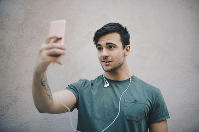 Young male computer programmer taking selfie with smart phone against beige wall in office