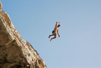 Low angle view of man jumping on rock against clear sky