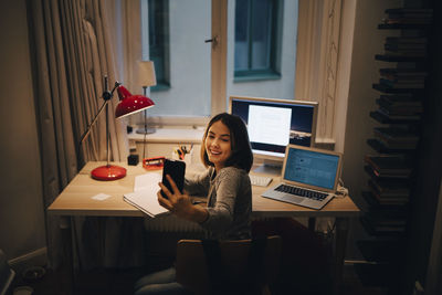 Girl taking selfie through smart phone while sitting at illuminated desk