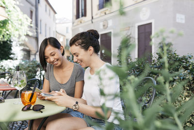 Italy, padua, two young women checking their cell phones at sidewalk cafe