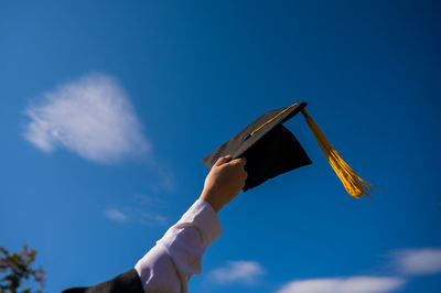 Cropped hand of woman holding mortarboard against blue sky