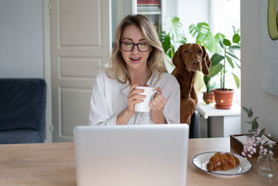 Woman in pajamas sitting with dog on chair at home drinking tea taking in video chat at laptop