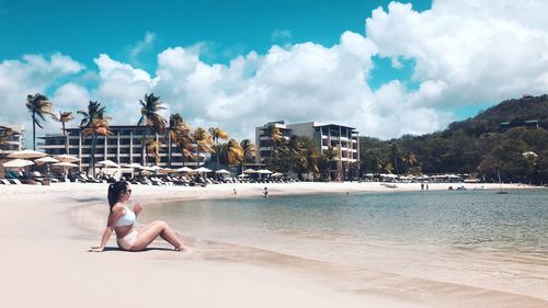 Side view of woman sitting at beach against cloudy sky