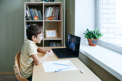 7 years old boy sitting by desk with laptop doing writing task during online lesson. side view