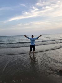 Rear view of man with arms outstretched standing at beach against sky