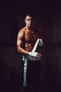 Portrait of shirtless male athlete wearing hand wrap while standing in darkroom