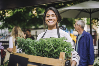 Young smiling female florist holding crate of plants at flea market