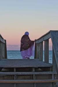 Rear view of man sitting on wood against sky during sunset