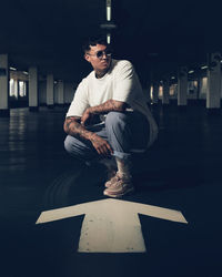 Young man wearing sunglasses crouching on arrow symbol in parking lot at night