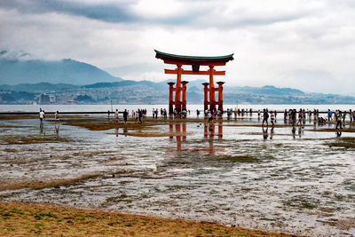 People at torii gate on shore against cloudy sky