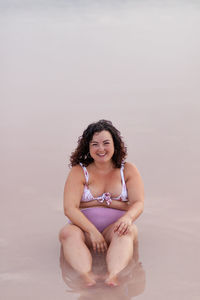Delighted curvy female in bikini sitting in water of pink pond in summer and looking at camera
