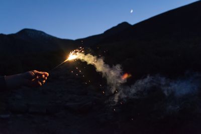 Cropped hand of person holding sparkler against mountain at night