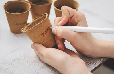 The hands of a caucasian girl hold a cardboard glass and sign the word tomato with a red marker