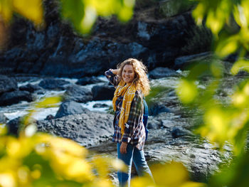 Young curly woman traveller looking at camera and smiling at mountain river in autumn