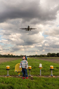 Full length of boy looking up with airplane flying over grass against sky