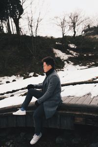 Young man sitting on snowy field against sky