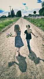 Rear view full length of sisters walking on dirt road amidst field