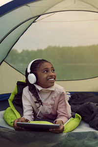 Smiling girl with headphones holding digital tablet while lying in tent