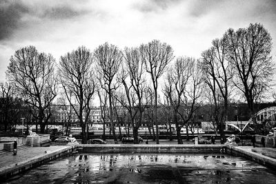 Bare trees by river against cloudy sky