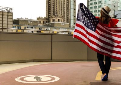 Rear view of woman walking on driveway while holding american flag against buildings