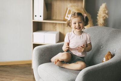 A little charming girl sits in a chair with her toy bear and smiles