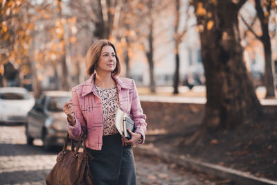 Stylish business mature woman 50-55 year old holding notebook and bag wearing suit walking outdoors.