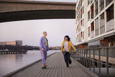 Female friends or gay couple on promenade, one woman running, while other skateboarding